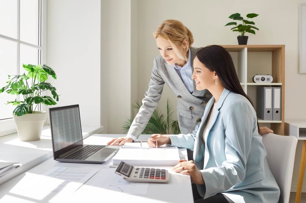 Two,Smiling,Businesswomen,Working,Together,In,Office,,Checking,Spreadsheet,On